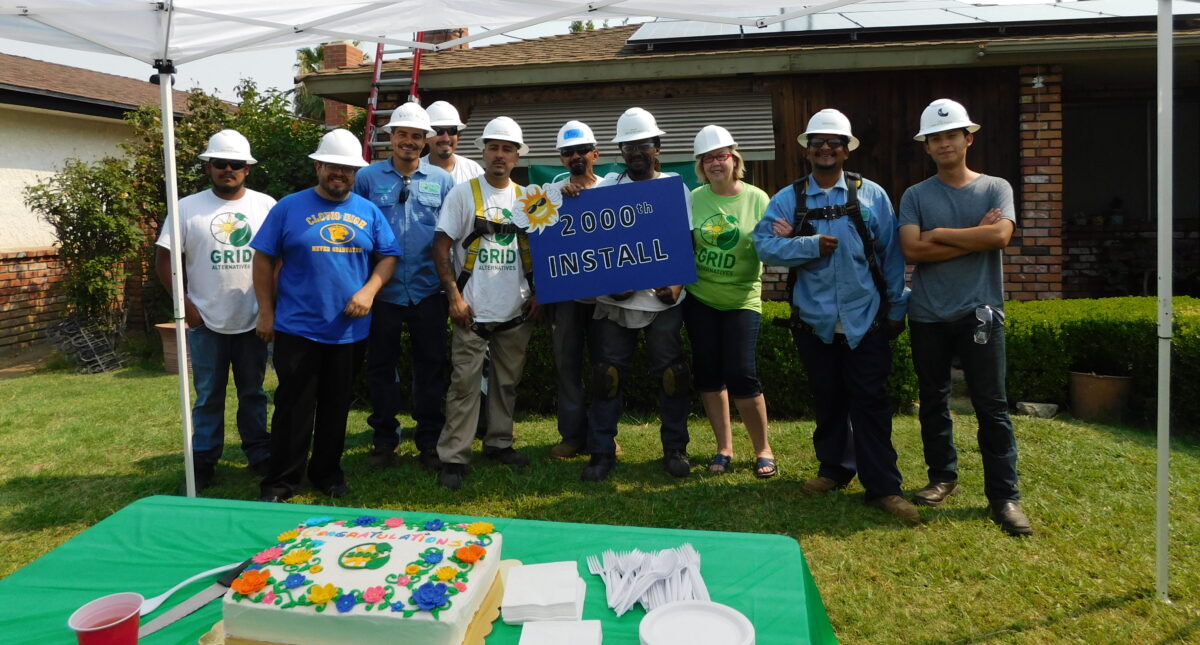 Team of workers in hard hats celebrating 1000th solar panel installation with cake and banner outdoors