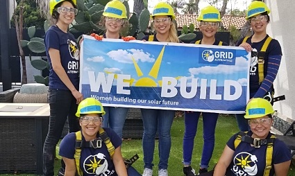 Seven students in hard hats and GRID Alternatives shirts holding a "We Build" banner outdoors