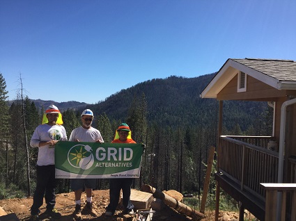Three people in safety gear hold a GRID Alternatives banner in a mountain forest setting near a cabin
