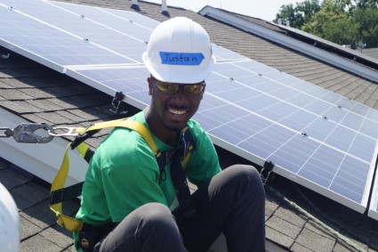 Worker in green shirt and white hard hat smiling on rooftop with solar panels behind