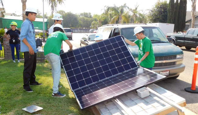 Group of workers in green shirts and hard hats demonstrating a large solar panel at an outdoor community event