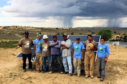 Ten people standing together outdoors on sandy ground with solar panels and stormy sky behind them