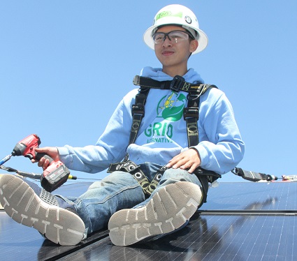 Worker in safety gear sitting on solar panels holding a power drill