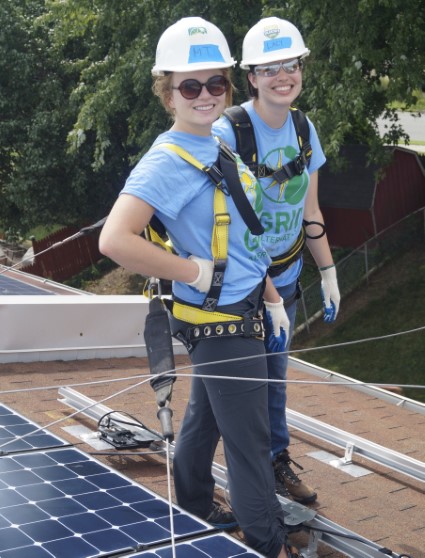 Two young women in safety harnesses and hard hats stand on solar panels, smiling at the camera