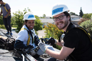 Two workers in hard hats and safety gear smile while installing solar panels on a residential rooftop