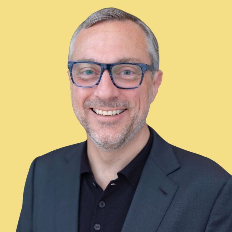 Professional headshot of man with glasses smiling against yellow background