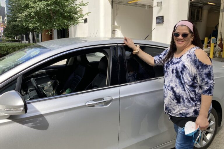 Woman in tie-dye shirt and pink visor posing beside open car door in parking garage