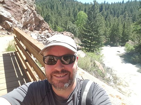 Man in white cap and sunglasses smiling at mountain scenic overlook with forest and river