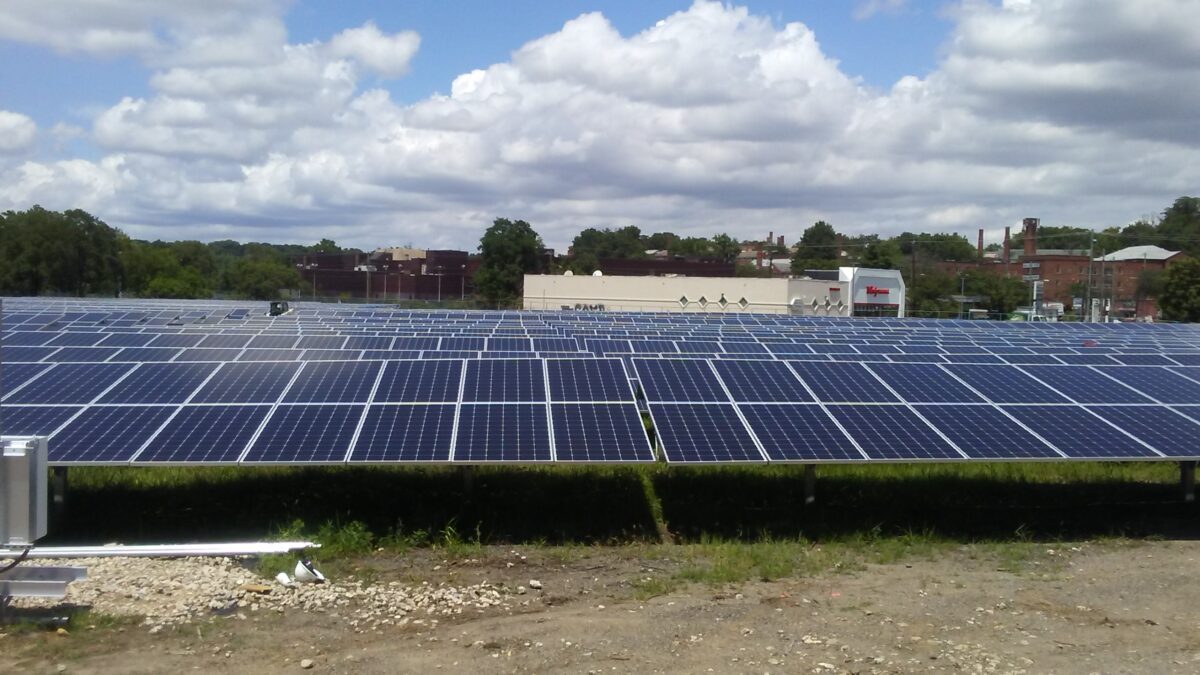 Large-scale solar panel array on rooftop with industrial buildings and trees in background under cloudy sky
