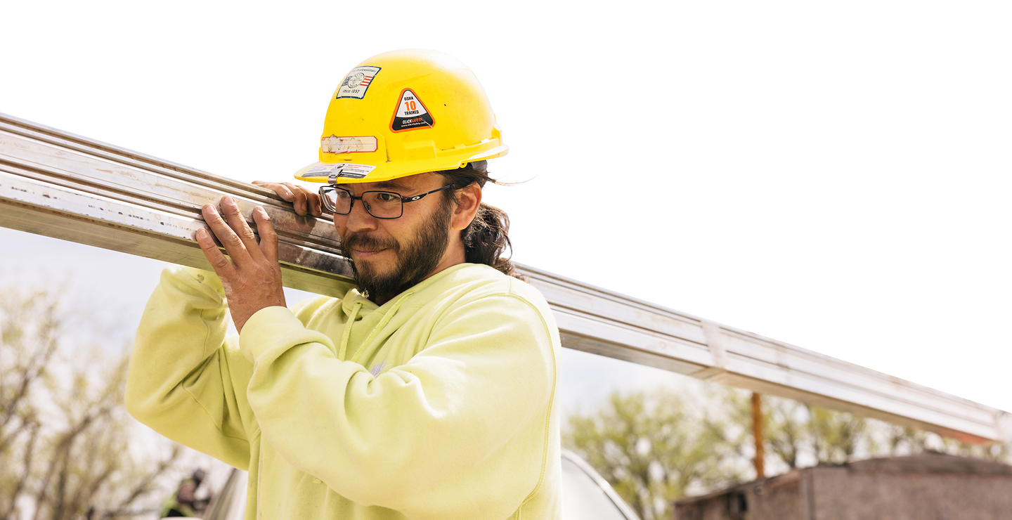 Construction worker in yellow safety helmet and high-visibility shirt carrying metal beam on shoulder