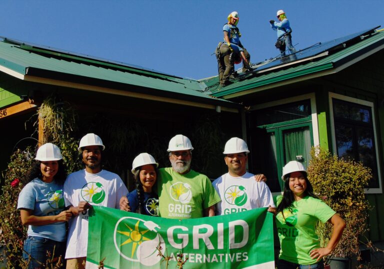 GRID Alternatives team holding banner in front of green building with workers on roof above