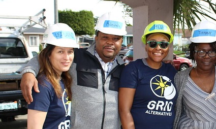 Four people wearing white hard hats and branded apparel stand together outdoors at what appears to be a community event or construction site visit