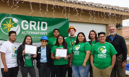 Group of people wearing green GRID Alternatives t-shirts posing together under a covered structure with icicle lights