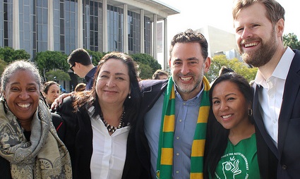 Five people smiling at outdoor event in front of building, one wearing green and gold scarf