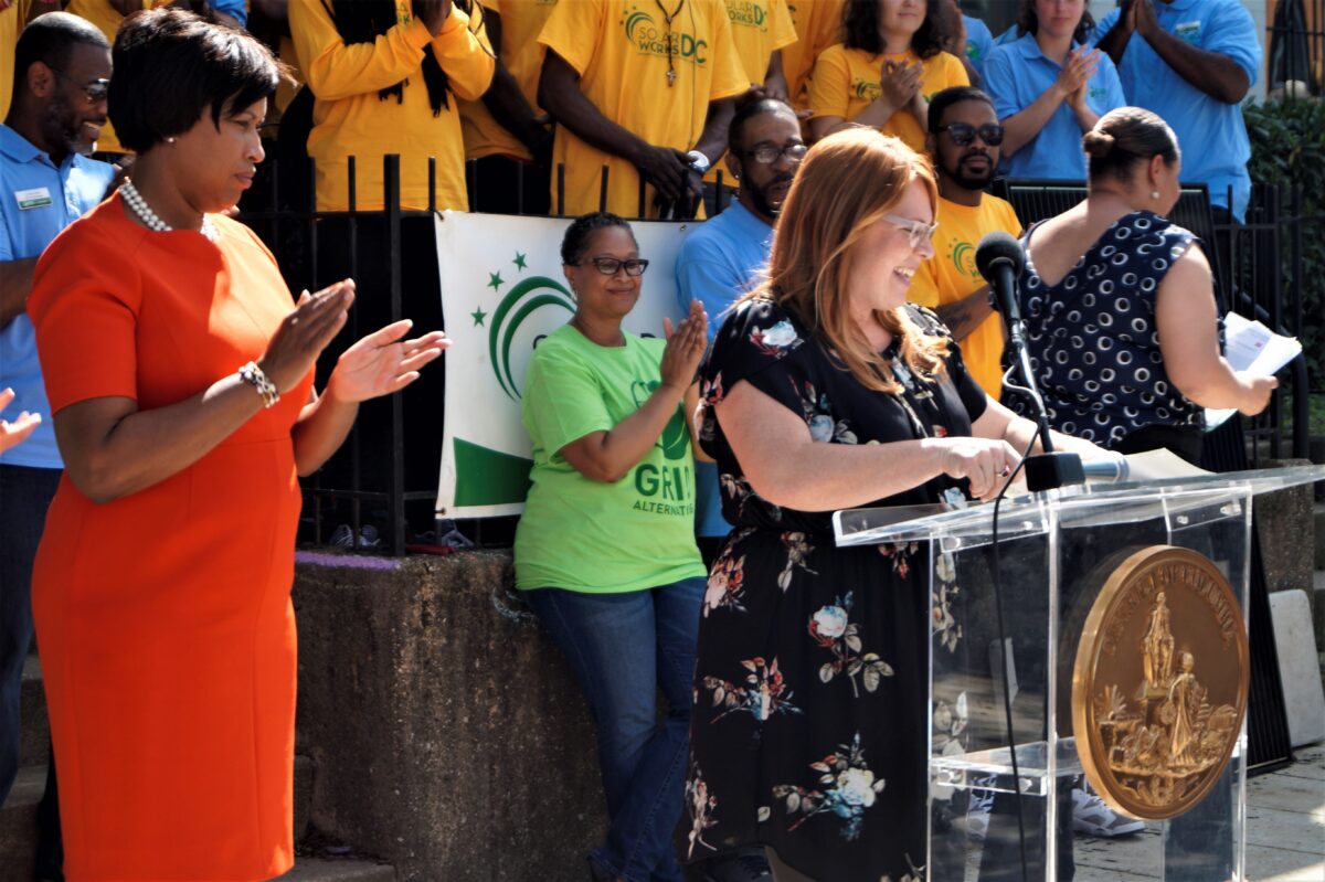 Woman speaking at podium during community event with supporters in yellow and green shirts behind her