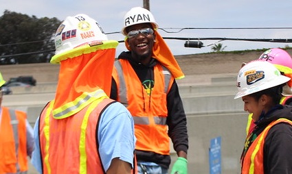 Construction workers in safety vests and hard hats smiling on job site, one wearing name tag "RYAN