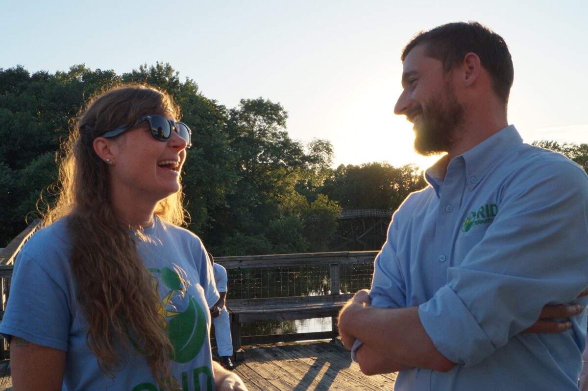 Two people smiling and conversing on a wooden bridge at sunset overlooking a river