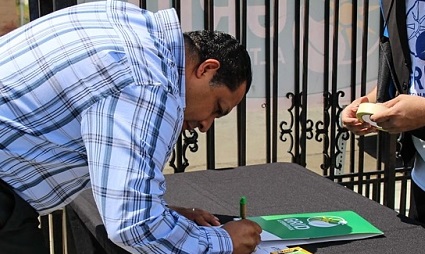 Man in blue plaid shirt signing green document at table with metal fence in background