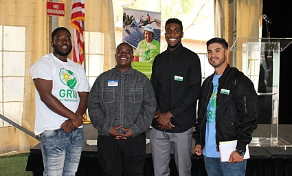 Four men smiling at camera in indoor venue, one wearing green shirt with logo, others in casual jackets and shirts
