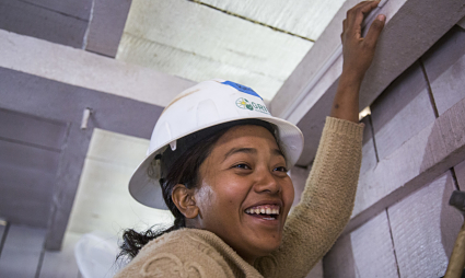 Smiling woman in hard hat reaching up while installing ceiling tiles