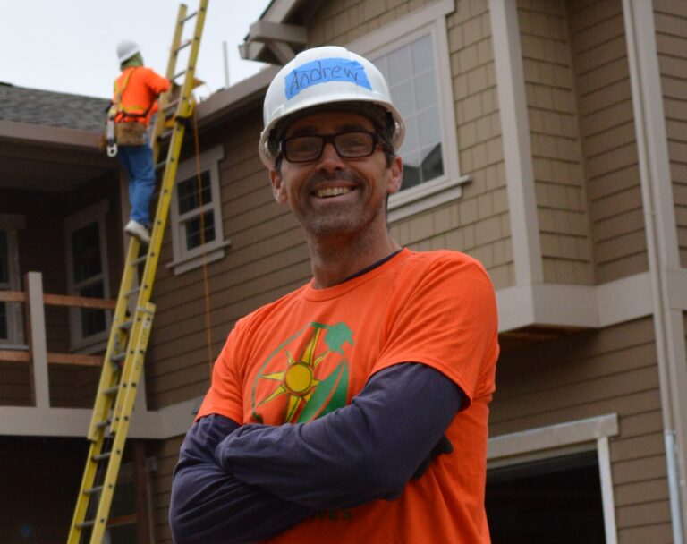 Man in hard hat and orange shirt smiling on construction site with ladder and worker behind him