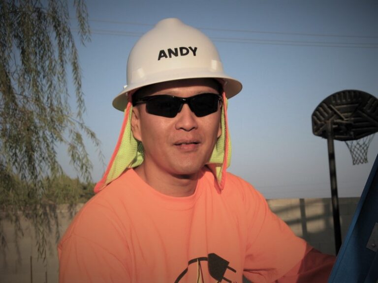 Person named Andy wearing white hard hat with neon yellow neck protector and orange shirt at outdoor construction site