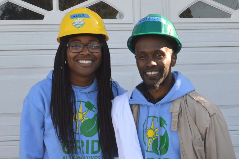 Two people wearing blue hoodies and construction hard hats smile in front of a white garage door