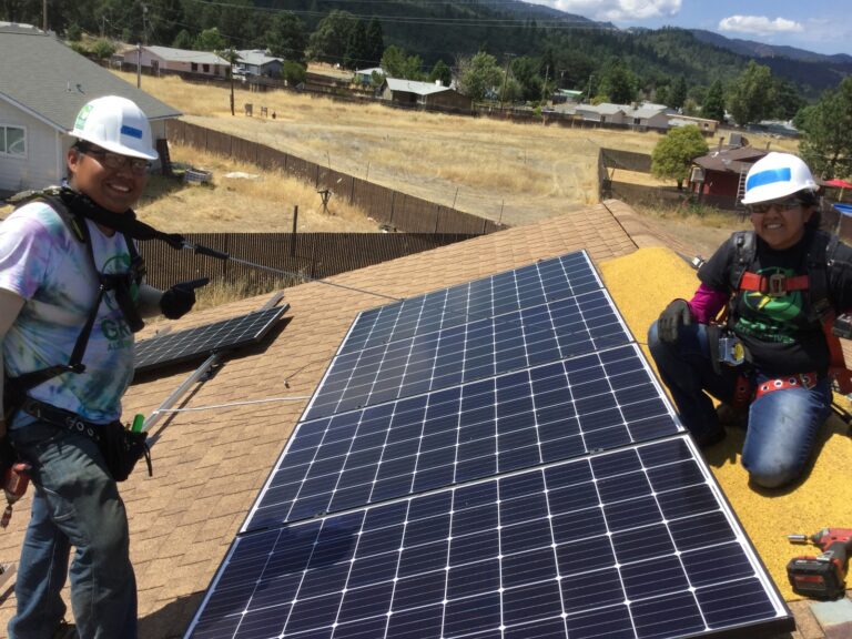 Two workers in safety gear installing solar panels on a residential roof in a mountain valley