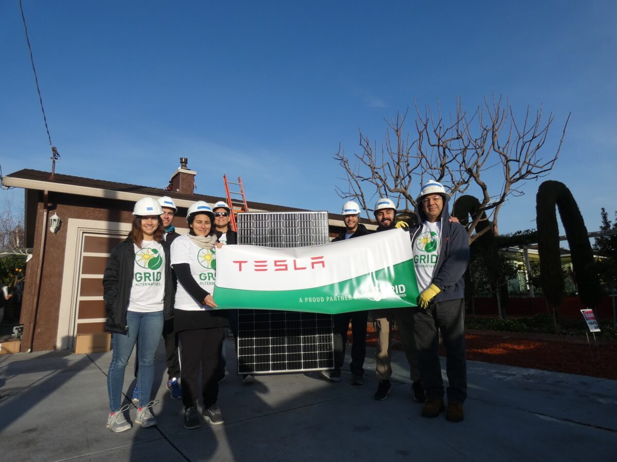 Team holding Tesla banner at solar panel installation site, wearing hard hats and safety gear
