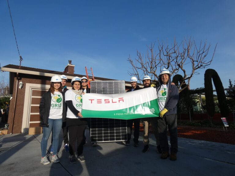 Team holding Tesla banner at solar panel installation site, wearing hard hats and safety gear