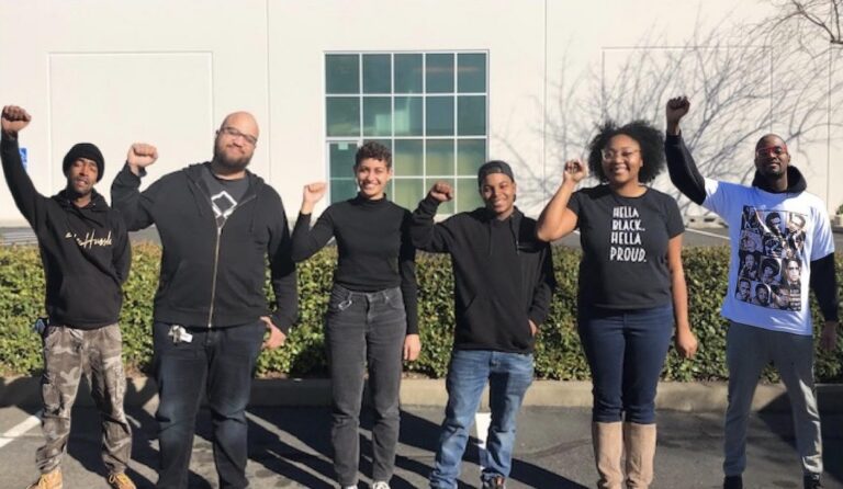 Six people raising fists in solidarity outdoors, one wearing "Hella Proud" shirt
