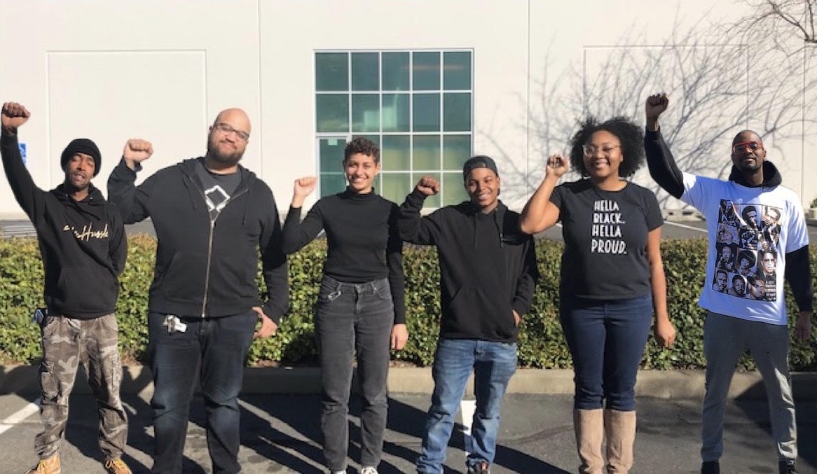 Six people raising fists in solidarity outdoors, one wearing "Hella Proud" shirt