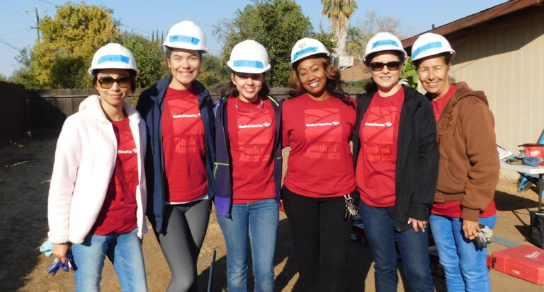 Six volunteers wearing red Bank of America t-shirts and white hard hats pose together at a construction site