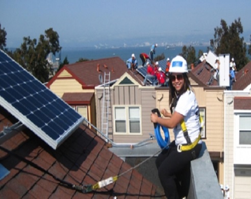 Woman wearing hard hat and safety gear on rooftop with solar panels and bay view