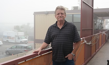 Man in black striped polo shirt standing on elevated rooftop platform overlooking parking area