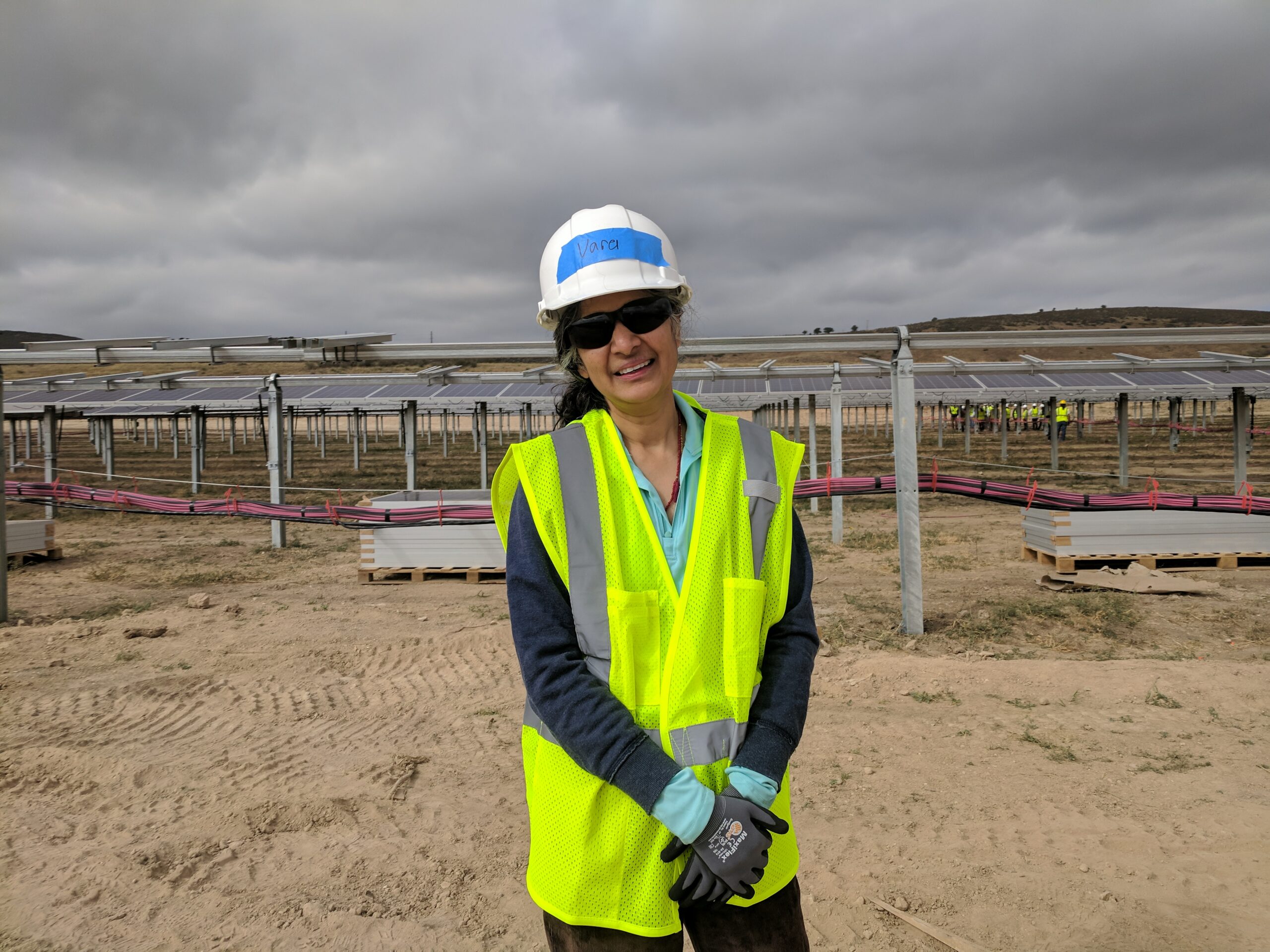 A woman smiles in front of a large community solar array, she is wearing hard hat, gloves, and safety vest