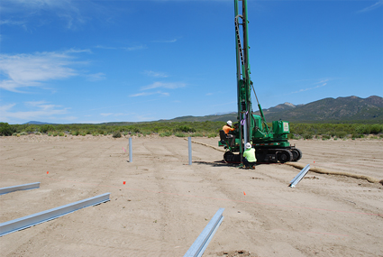 Green drilling rig on sandy desert ground with blue PVC pipes and mountains in background