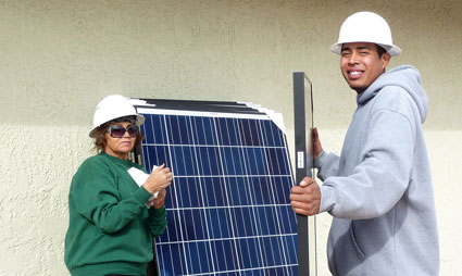 Two workers in hard hats holding a solar panel against a building wall