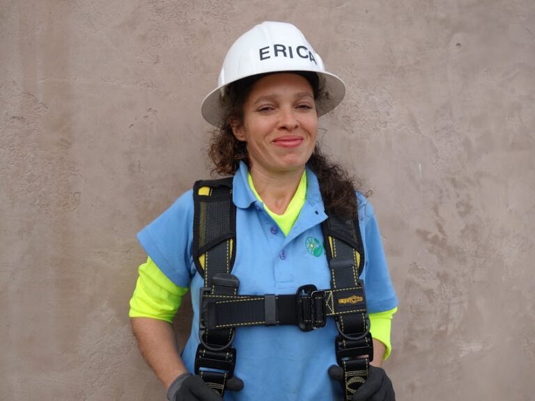 Woman in safety gear with "ERICH" hard hat and harness smiling at camera