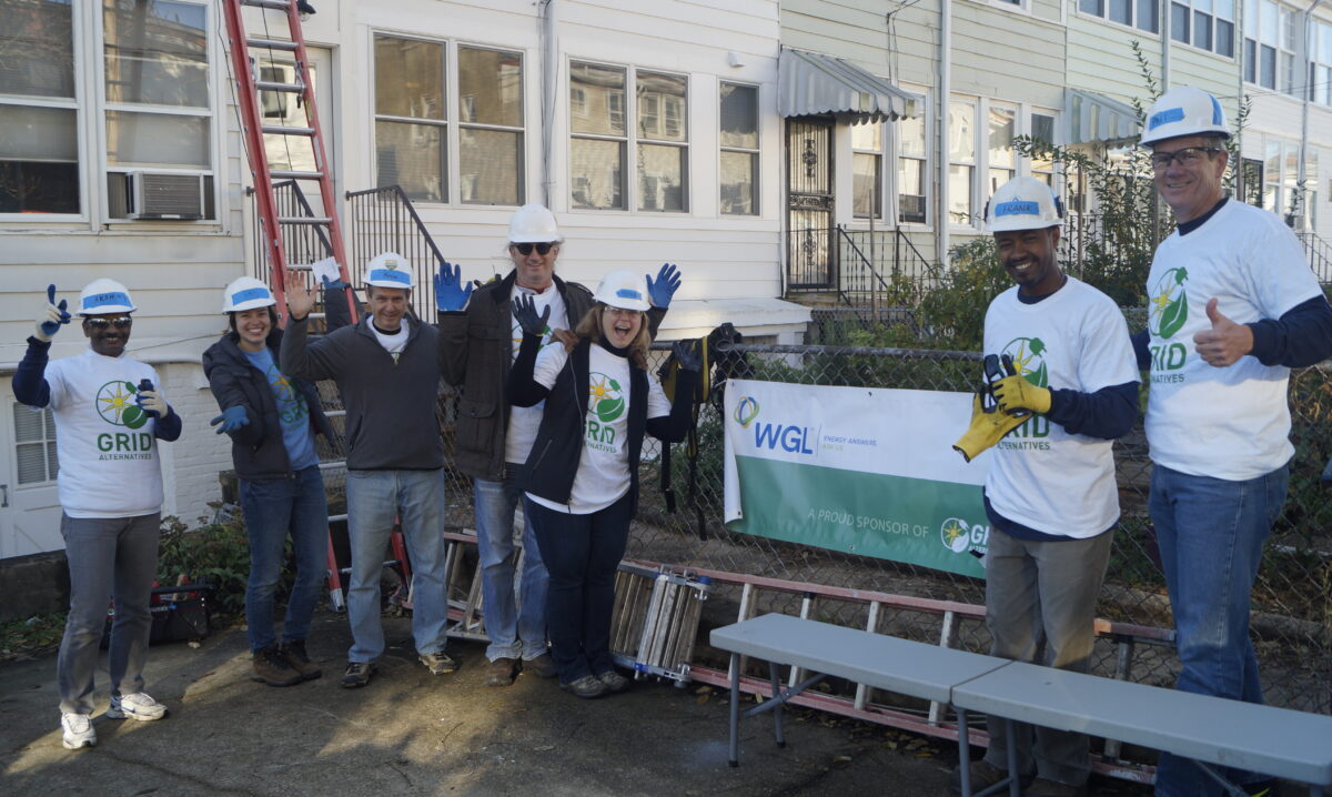 Community volunteers in hard hats and GRID t-shirts hold WGL sponsor banner at outdoor worksite