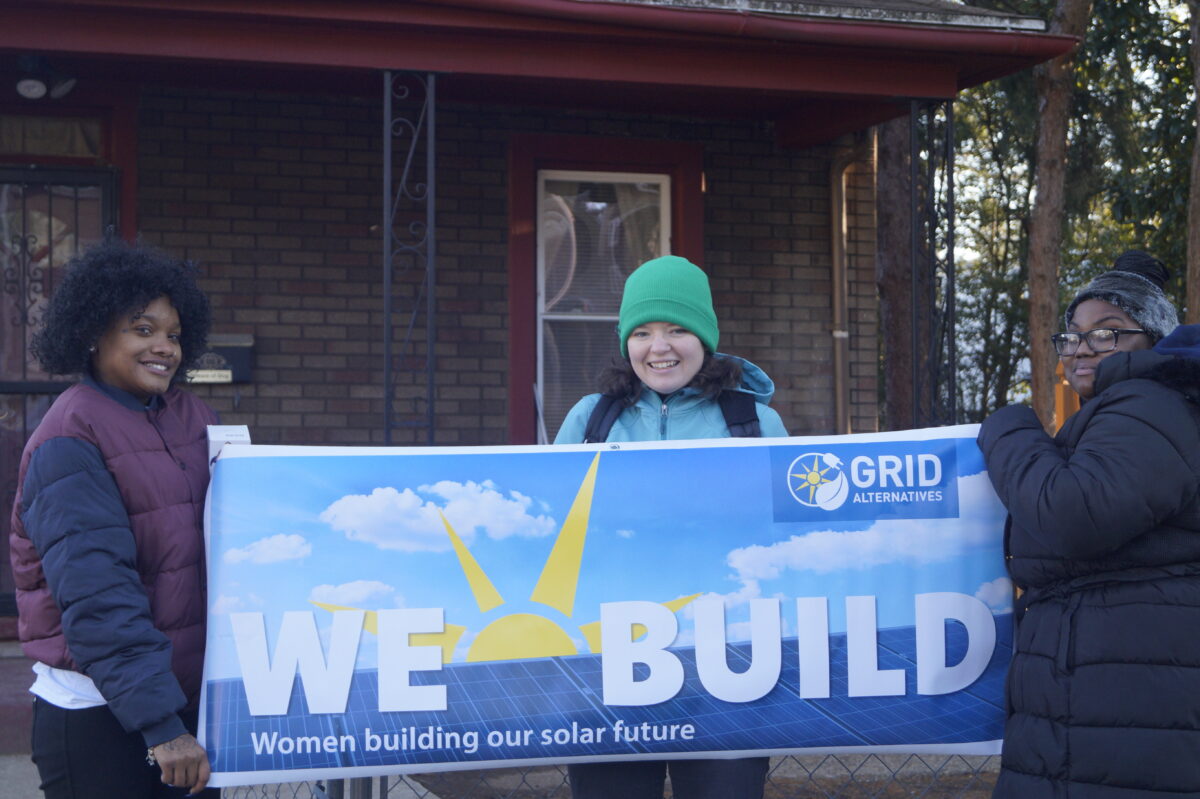 Three women holding a "We Build" banner for GRID Alternatives' solar energy program outside a brick building