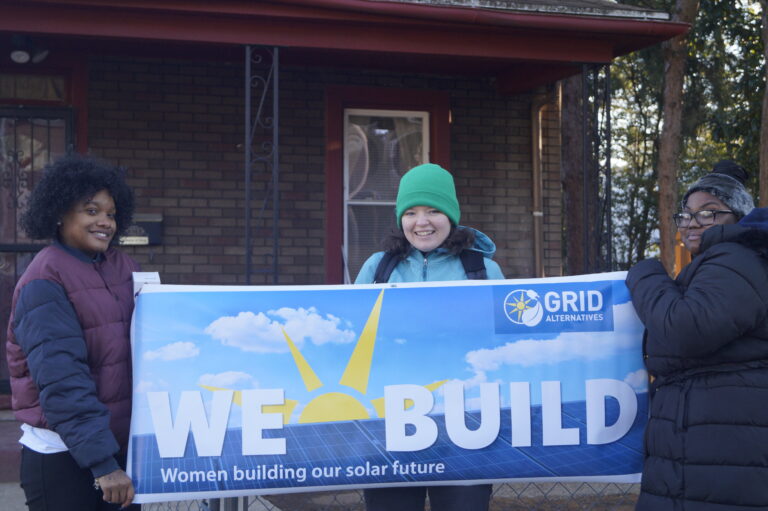 Three women holding a "We Build" banner for GRID Alternatives' solar energy program outside a brick building