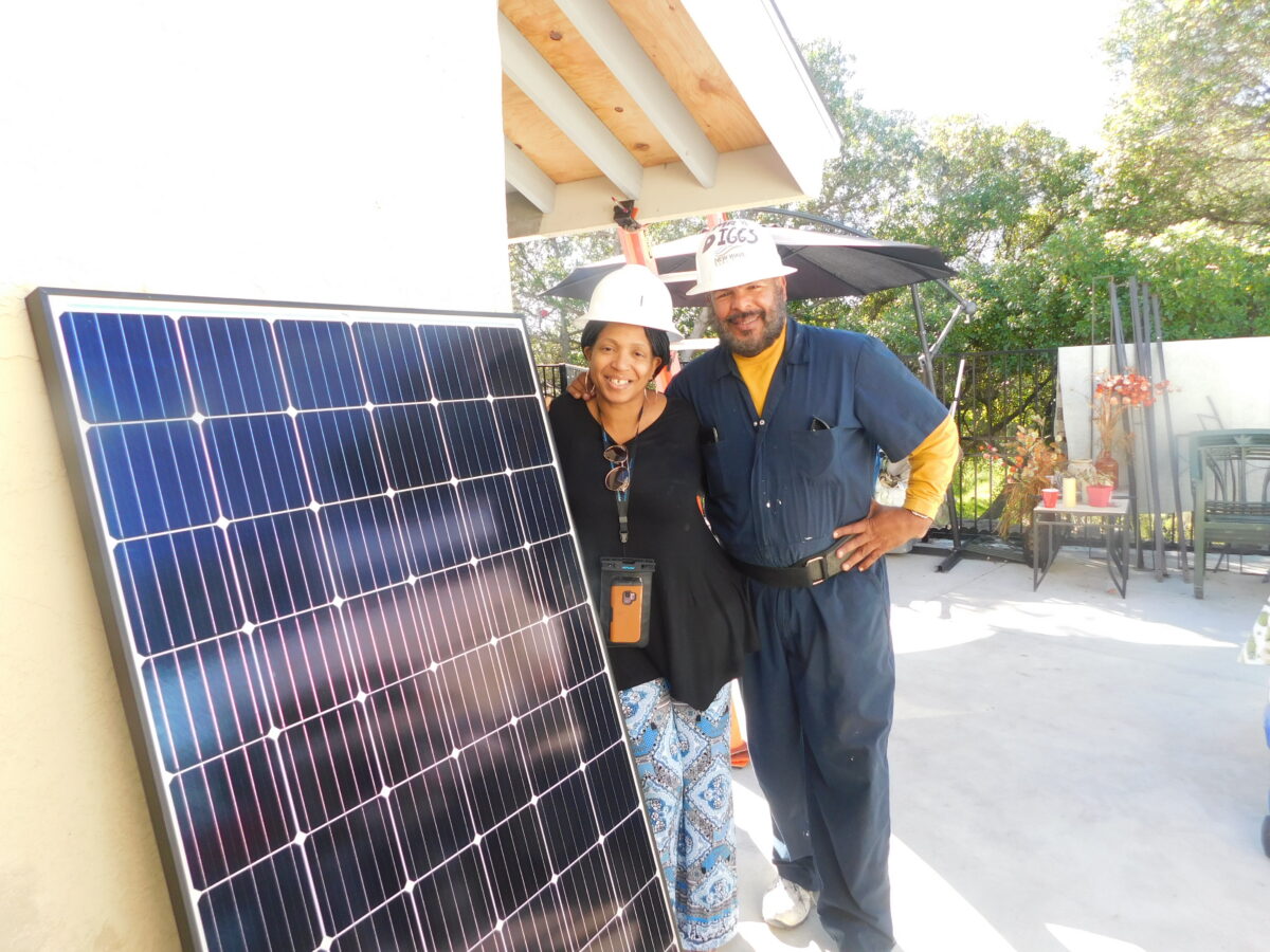 Two workers in hard hats stand beside a large blue solar panel on a residential rooftop