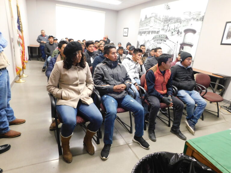 Group of people seated in a community meeting room with an American flag and Fresno historical poster visible