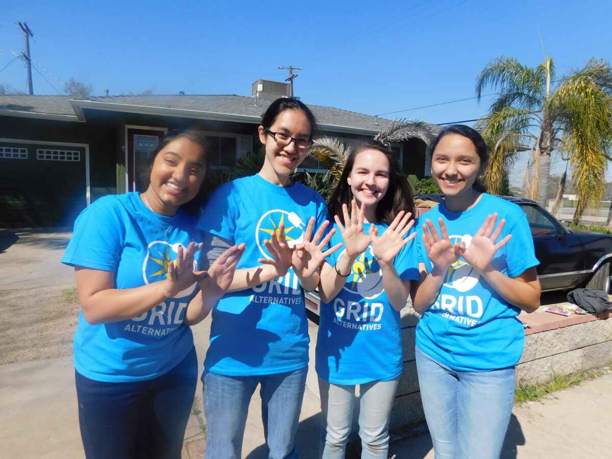Four smiling teenage girls in bright blue "Grid Alternatives" t-shirts waving at camera in front of a house
