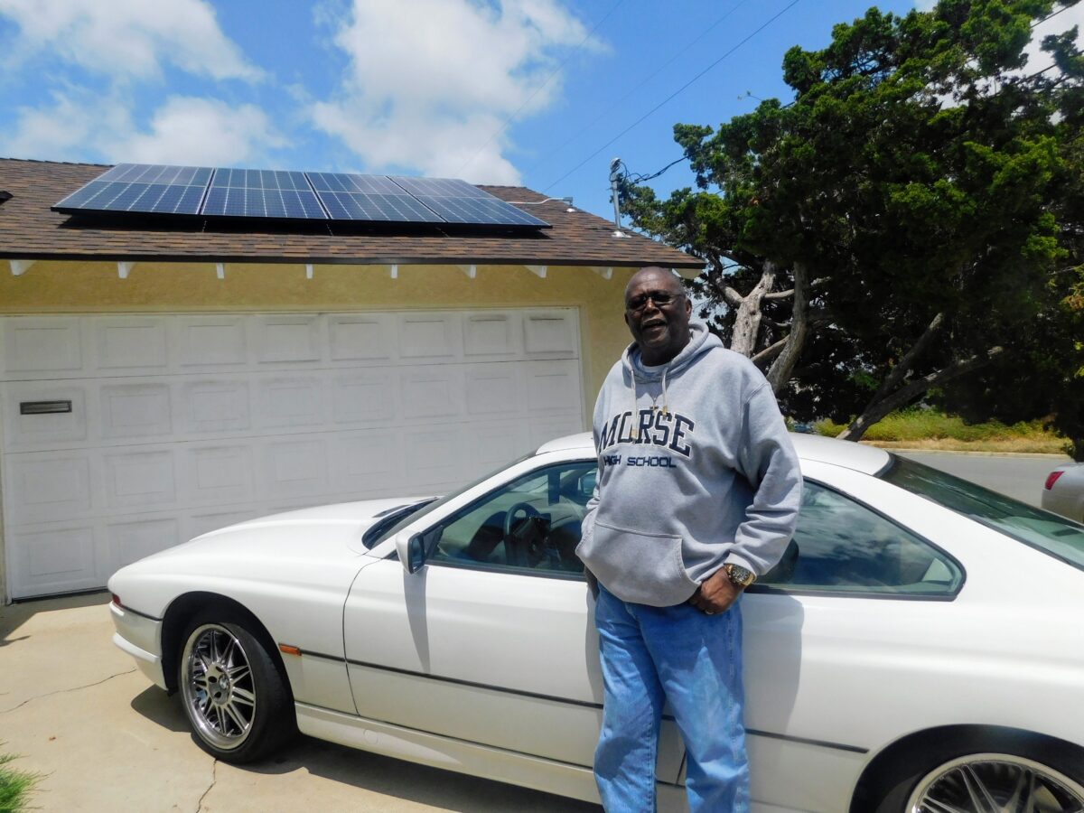 Man in "Morse High School" hoodie standing beside white coupe in driveway with solar panels on garage roof