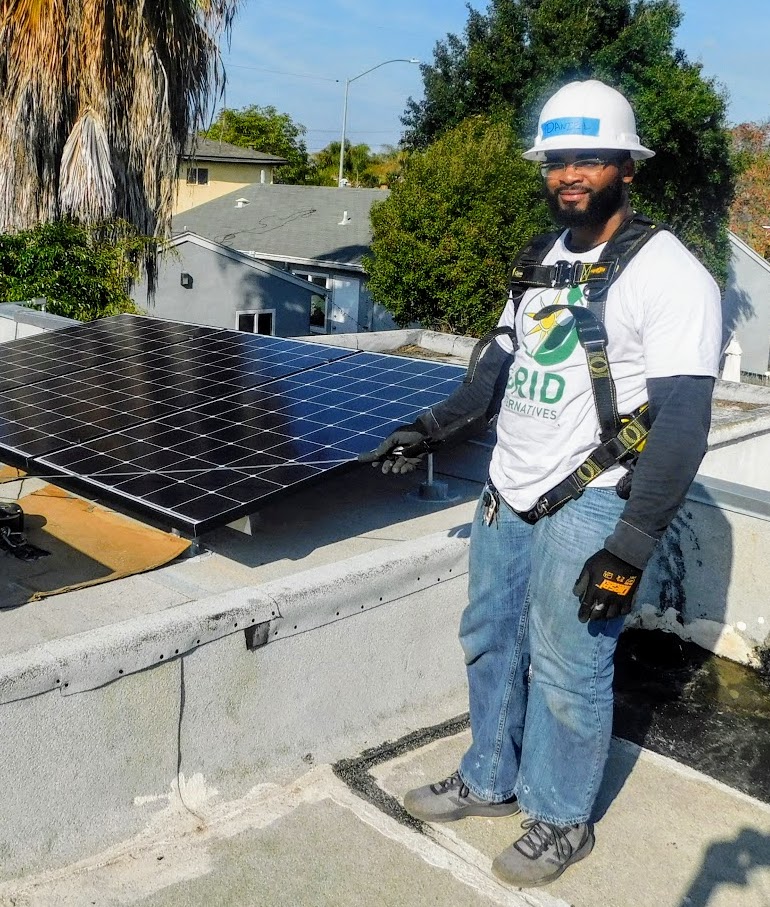 Solar technician in safety gear standing next to rooftop solar panels at residential installation