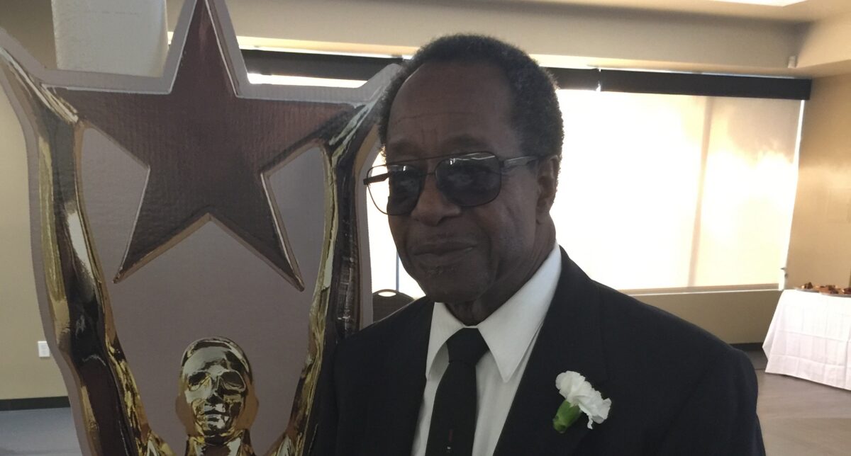 Man in sunglasses and formal attire with white flower boutonniere next to award statue