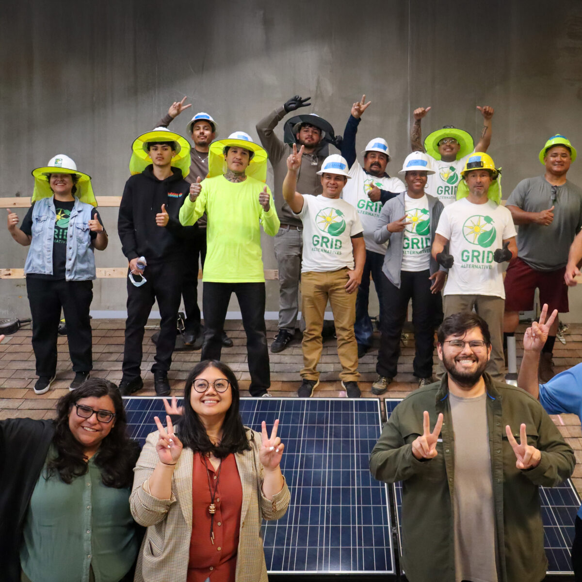 A large group of IBT trainees and instructors celebrate together around a solar panel at the end of a cohort.