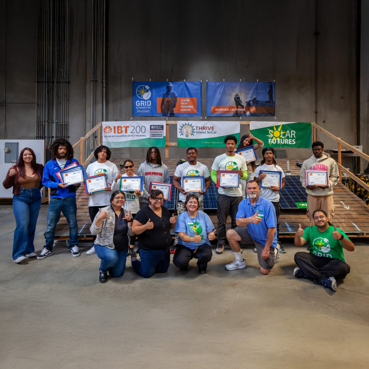 Graduating IBT 200 trainees stand together holding their certificates in front of a GRID Alternatives banner.